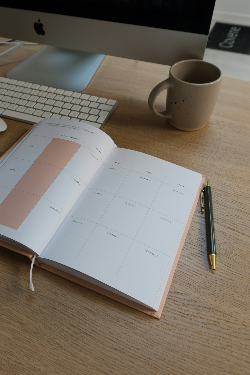 Schreibtisch mit offenem Planer, Stift, Tasse und Computer.