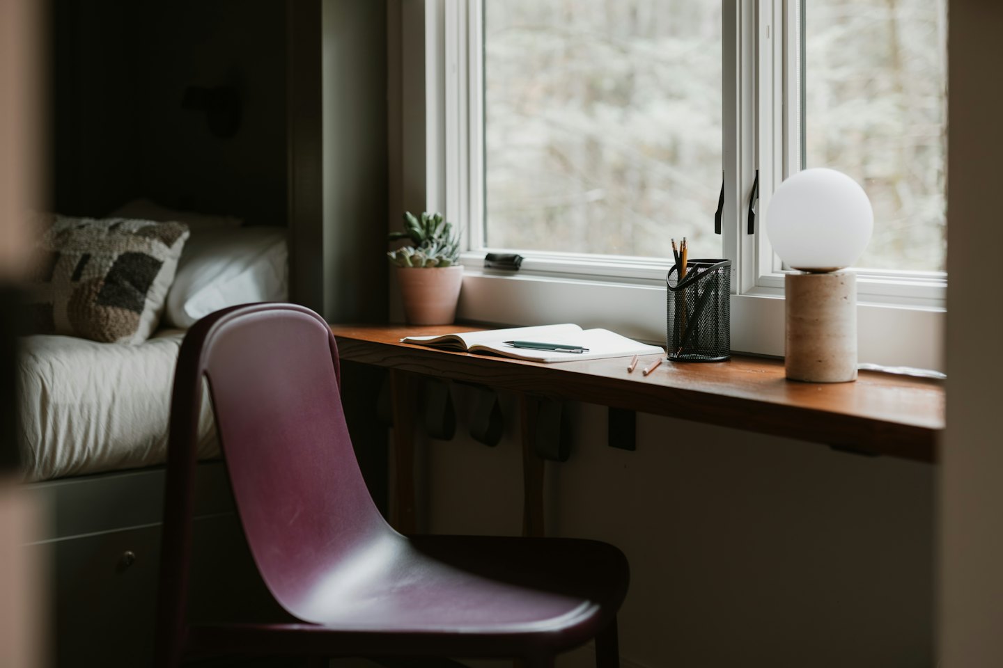 Schreibtisch mit Notizbuch und Lampe am Fenster mit Blick auf Bäume.